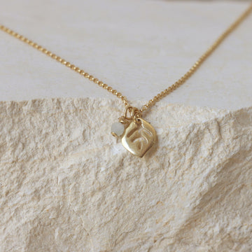 the gold-plated leaf pendant and moonstone charm placed on a light travertine/marble background. Soft, creamy tones complement the warm gold and the opalescent moonstone.
